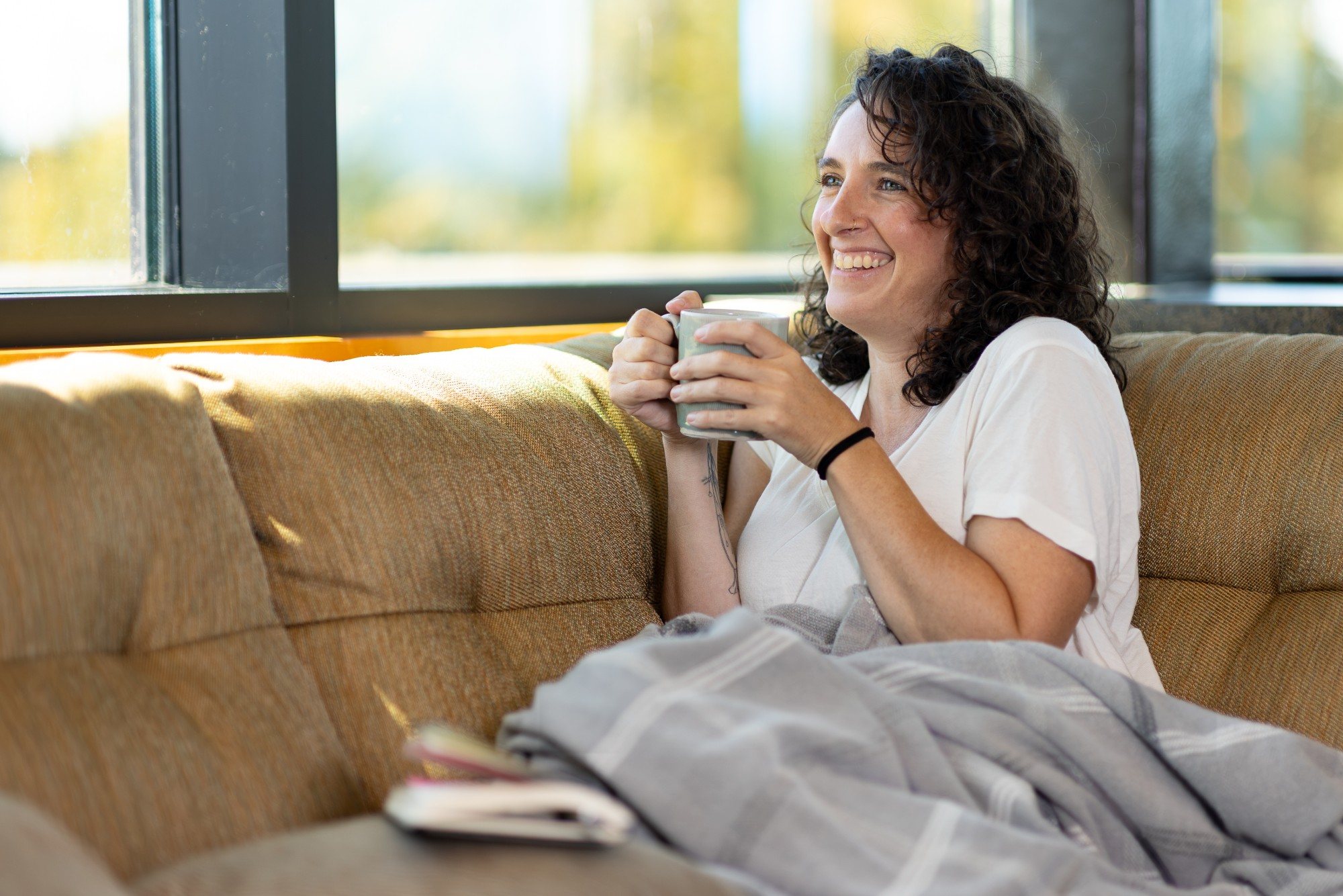 A smiling woman holding a mug on a sofa by a sunny window
